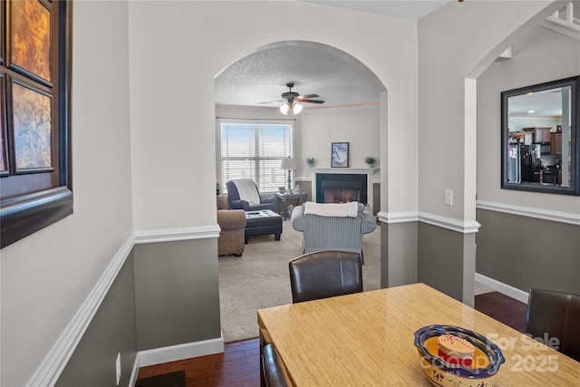 dining space with a textured ceiling, ceiling fan, baseboards, a lit fireplace, and dark wood-style floors