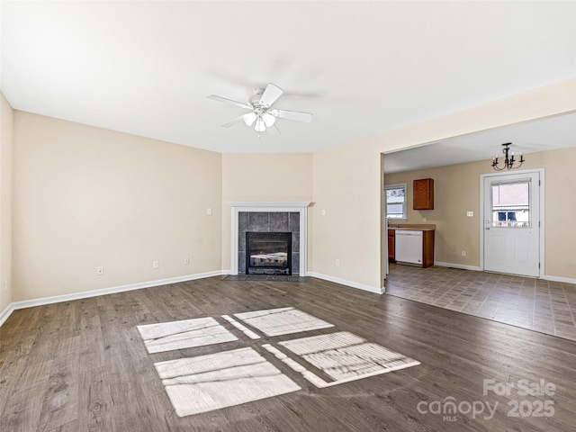 unfurnished living room with baseboards, dark wood finished floors, a tiled fireplace, and ceiling fan with notable chandelier