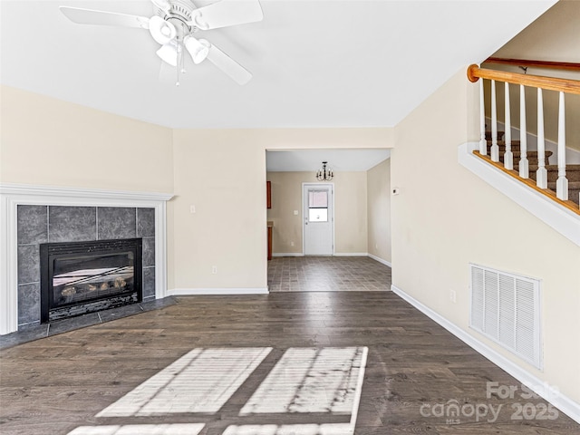 unfurnished living room featuring dark wood-style floors, visible vents, a tiled fireplace, baseboards, and ceiling fan with notable chandelier