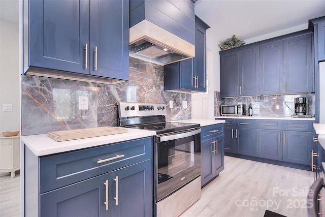 kitchen featuring blue cabinetry, custom exhaust hood, light wood finished floors, light countertops, and appliances with stainless steel finishes