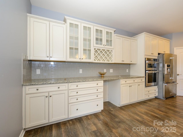 kitchen with light stone counters, tasteful backsplash, dark wood finished floors, stainless steel appliances, and glass insert cabinets