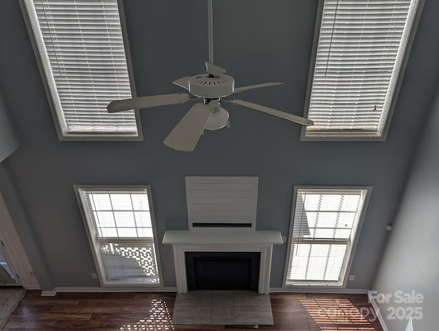 unfurnished living room featuring ceiling fan, a fireplace, wood finished floors, a towering ceiling, and baseboards