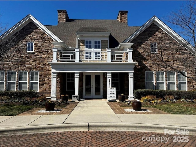 view of front facade featuring french doors, stone siding, a balcony, and a chimney