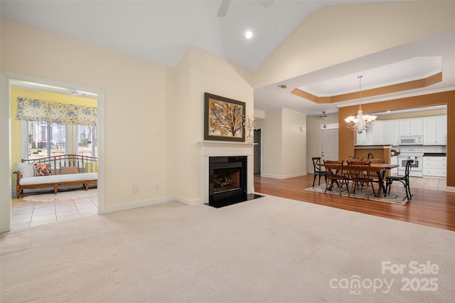 living area with baseboards, a fireplace with flush hearth, a tray ceiling, light carpet, and ceiling fan with notable chandelier