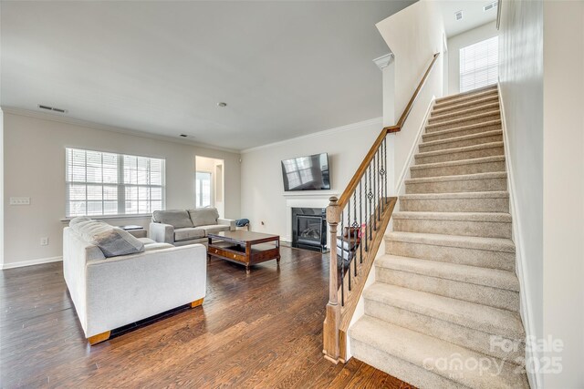 living room featuring dark wood finished floors, visible vents, stairway, ornamental molding, and a glass covered fireplace