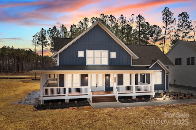 back of house at dusk with covered porch, a yard, board and batten siding, and roof with shingles