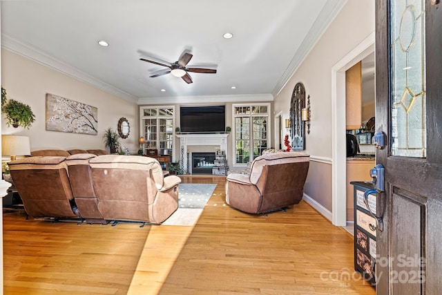 living room with ceiling fan, ornamental molding, a glass covered fireplace, and light wood-style flooring