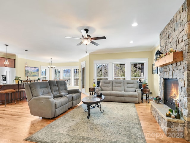 living room with light wood-style flooring, ceiling fan with notable chandelier, ornamental molding, and a stone fireplace