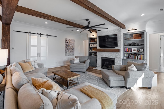 living area featuring a barn door, light wood-style flooring, a large fireplace, a ceiling fan, and beamed ceiling