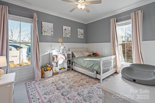 bedroom featuring light colored carpet, a decorative wall, ornamental molding, a ceiling fan, and wainscoting