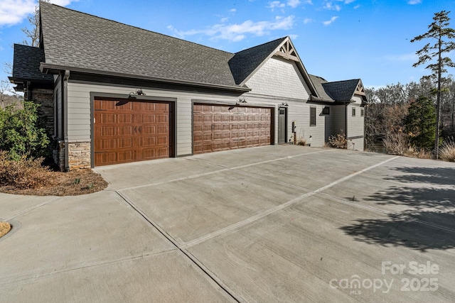 view of side of property with a garage, concrete driveway, a shingled roof, and stone siding