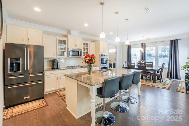 kitchen featuring a center island with sink, appliances with stainless steel finishes, glass insert cabinets, a kitchen breakfast bar, and decorative light fixtures