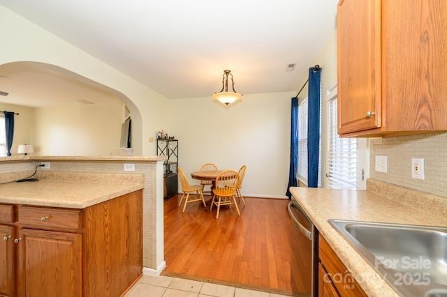 kitchen featuring light countertops, stainless steel dishwasher, decorative backsplash, brown cabinetry, and pendant lighting