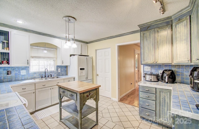 kitchen with a sink, ornamental molding, tile counters, white fridge with ice dispenser, and pendant lighting