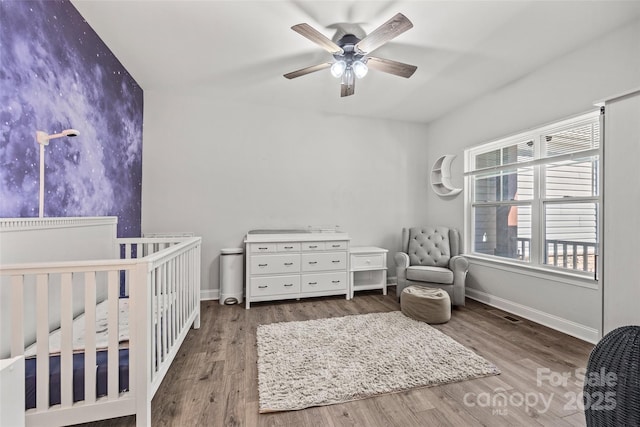 bedroom with a nursery area, ceiling fan, baseboards, and wood finished floors