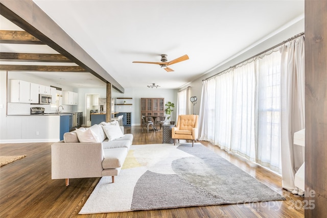 living area with dark wood-style flooring, beamed ceiling, a ceiling fan, and baseboards