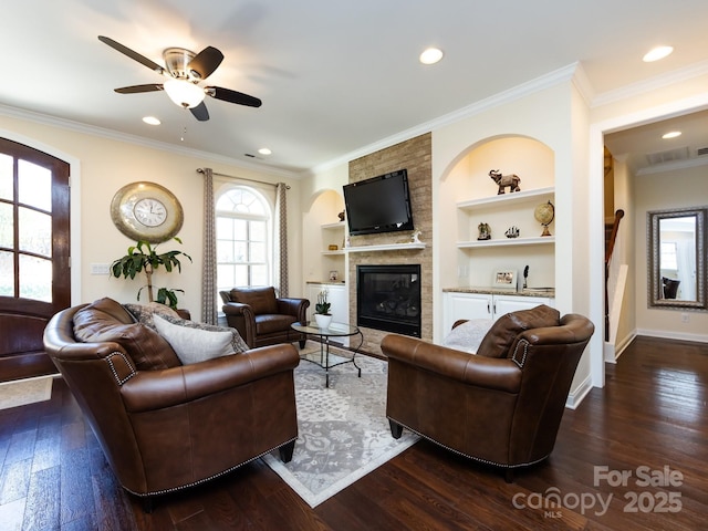 living area with built in features, dark wood finished floors, a ceiling fan, crown molding, and a fireplace