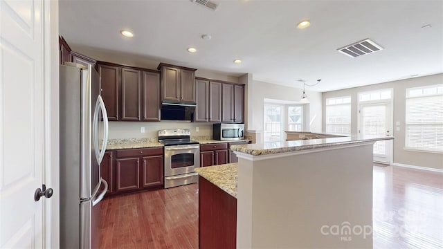 kitchen with light stone counters, a kitchen island with sink, under cabinet range hood, visible vents, and appliances with stainless steel finishes