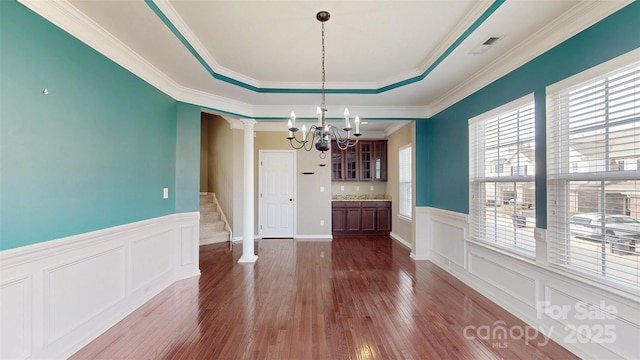 unfurnished dining area with a tray ceiling, decorative columns, stairway, dark wood-type flooring, and a chandelier