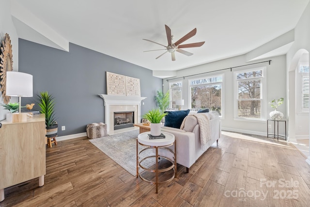 living area featuring wood finished floors, baseboards, a fireplace, arched walkways, and ceiling fan