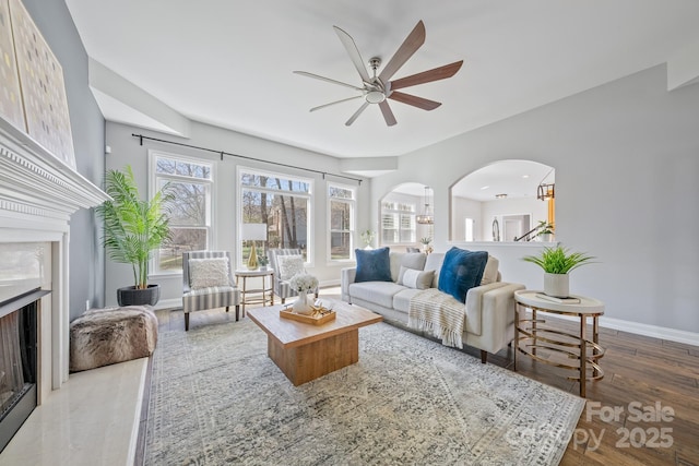 living room featuring arched walkways, a fireplace, wood finished floors, a ceiling fan, and baseboards