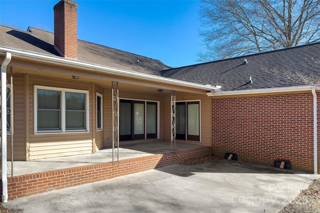 rear view of property featuring brick siding, a chimney, a patio area, and a shingled roof