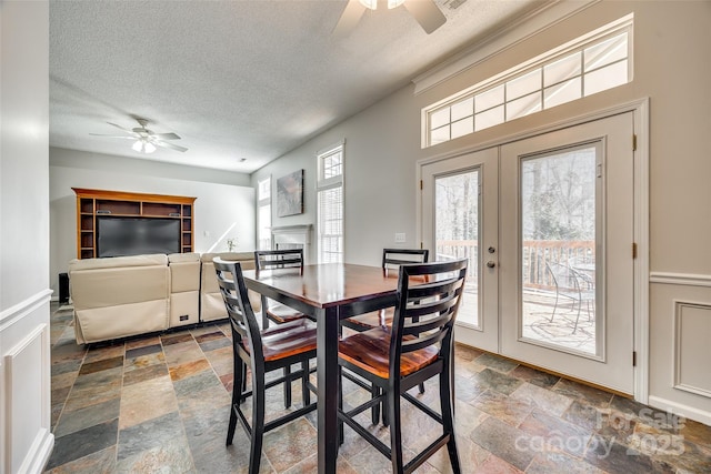 dining area with a textured ceiling, ceiling fan, a fireplace, french doors, and stone tile flooring