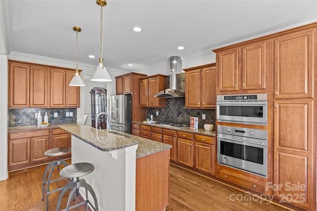 kitchen featuring dark wood finished floors, stainless steel appliances, brown cabinetry, a sink, and wall chimney exhaust hood