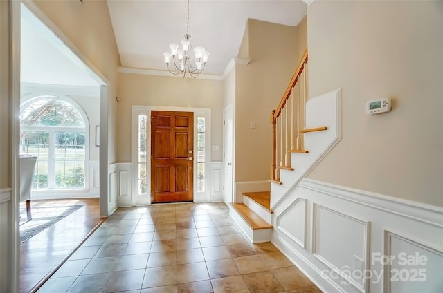 entryway with a wainscoted wall, an inviting chandelier, stairs, crown molding, and light tile patterned flooring