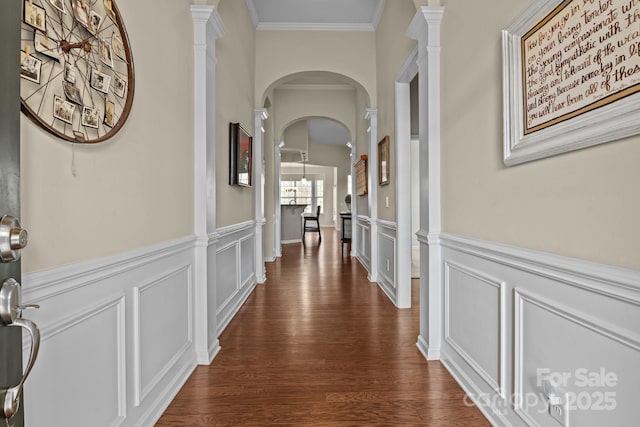 hall with arched walkways, a wainscoted wall, ornamental molding, dark wood-type flooring, and a decorative wall