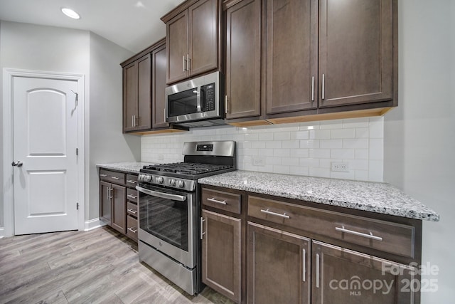 kitchen featuring dark brown cabinetry, stainless steel appliances, light wood-style floors, decorative backsplash, and light stone countertops