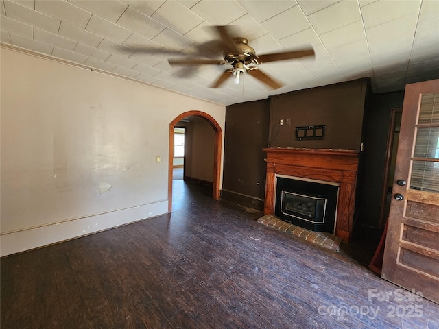 unfurnished living room featuring arched walkways, dark wood-style flooring, a ceiling fan, a brick fireplace, and baseboards