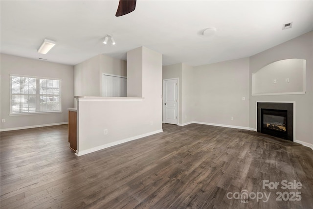 unfurnished living room featuring dark wood-style flooring, visible vents, a glass covered fireplace, ceiling fan, and baseboards