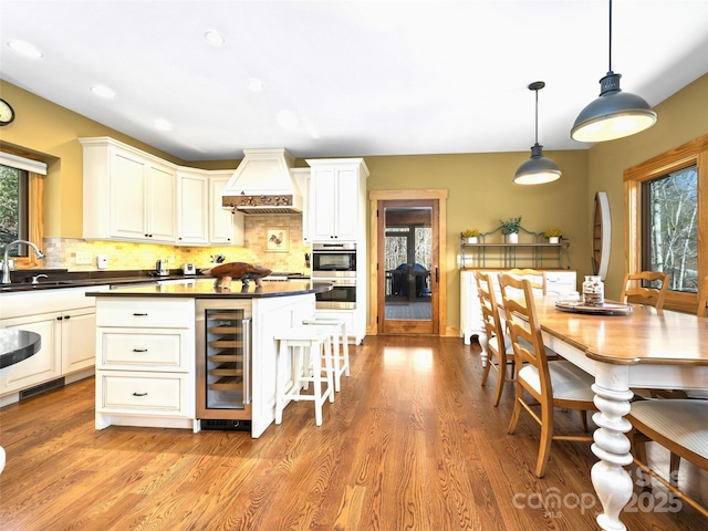 kitchen with pendant lighting, custom exhaust hood, dark countertops, a sink, and beverage cooler