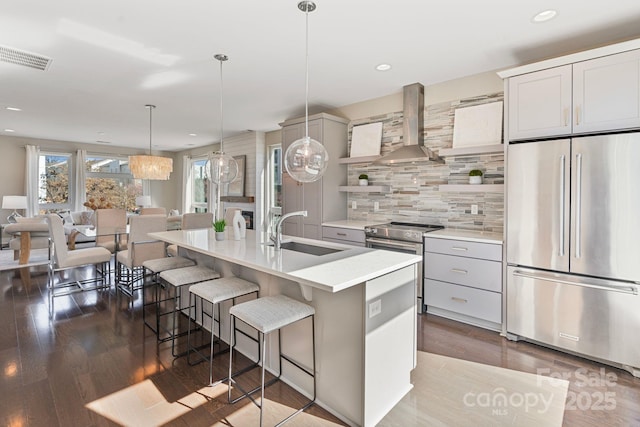 kitchen featuring open shelves, light countertops, appliances with stainless steel finishes, a sink, and wall chimney exhaust hood