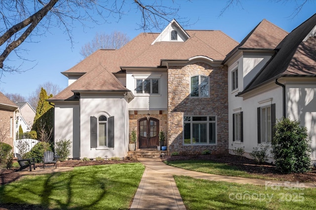 french country inspired facade with a front yard, stone siding, roof with shingles, and stucco siding