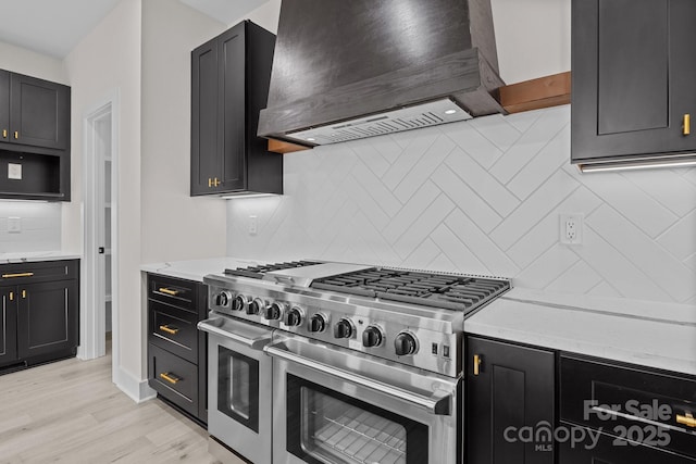 kitchen with decorative backsplash, custom range hood, dark cabinets, light stone countertops, and double oven range