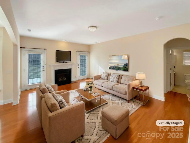 living room featuring arched walkways, a fireplace with flush hearth, wood finished floors, and baseboards