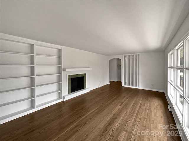 unfurnished living room featuring built in features, arched walkways, a tiled fireplace, dark wood-type flooring, and baseboards