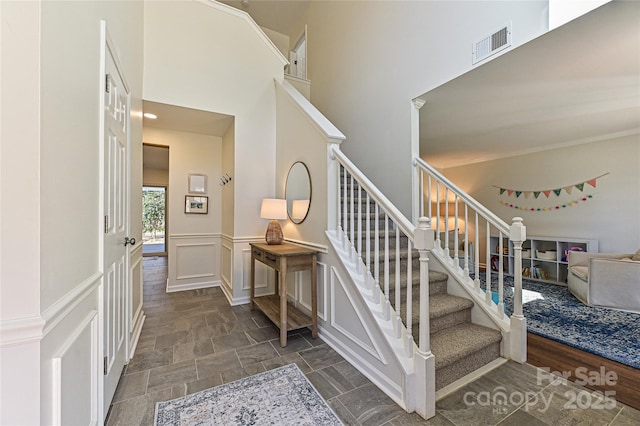 stairs with visible vents, a decorative wall, a high ceiling, stone finish floor, and wainscoting