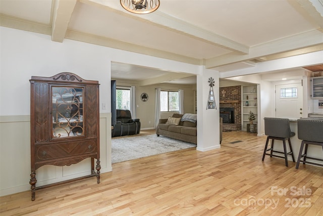 living area featuring baseboards, light wood-type flooring, a brick fireplace, built in shelves, and beam ceiling