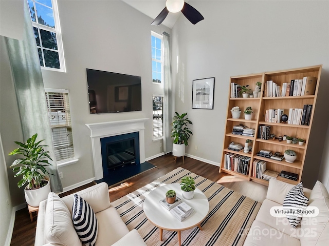 living room featuring baseboards, a fireplace with flush hearth, ceiling fan, wood finished floors, and a high ceiling