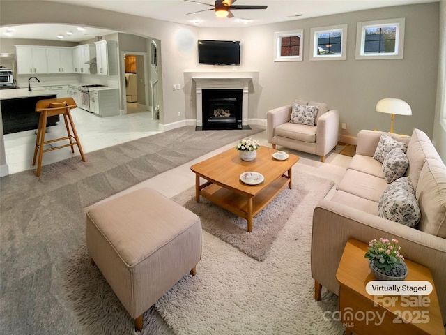 living area featuring arched walkways, a fireplace, light colored carpet, ceiling fan, and baseboards