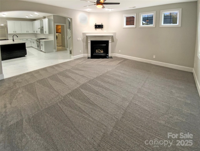 unfurnished living room with arched walkways, light colored carpet, a fireplace with flush hearth, ceiling fan, and baseboards