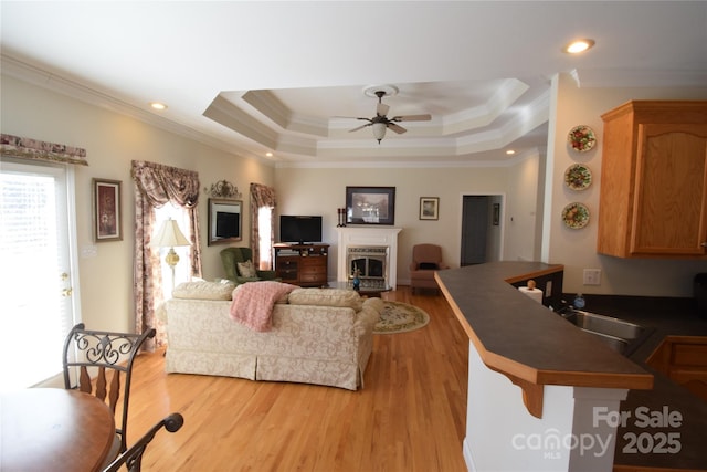 living room featuring a raised ceiling, ceiling fan, ornamental molding, light wood-style floors, and a fireplace