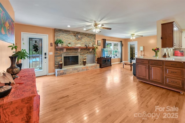 living area with light wood-type flooring, ceiling fan, a stone fireplace, and baseboards