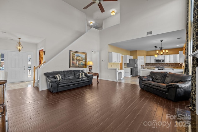 living room with stairs, visible vents, a towering ceiling, wood finished floors, and ceiling fan with notable chandelier