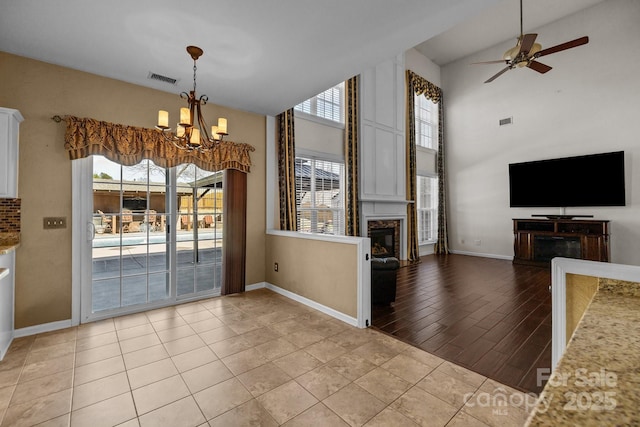 unfurnished dining area with baseboards, visible vents, a glass covered fireplace, high vaulted ceiling, and ceiling fan with notable chandelier