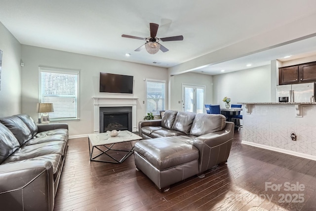 living area featuring ceiling fan, baseboards, dark wood finished floors, and a glass covered fireplace