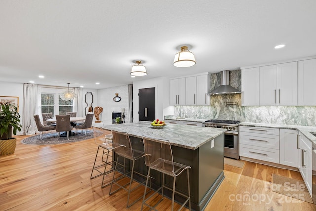 kitchen with light wood finished floors, a center island, stainless steel stove, wall chimney range hood, and a kitchen bar